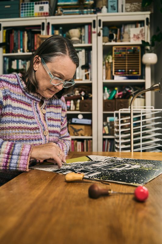 A printmaker carving a lino block at her desk, carving tools beside her