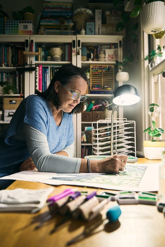 A printmaker in a blue knit vest sketching at a wooden desk in her home studio, bookshelves behind her