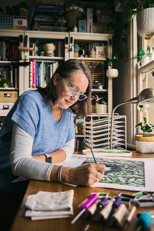 A printmaker sketching a leafy design, her carving tools and brushes laid out on the desk