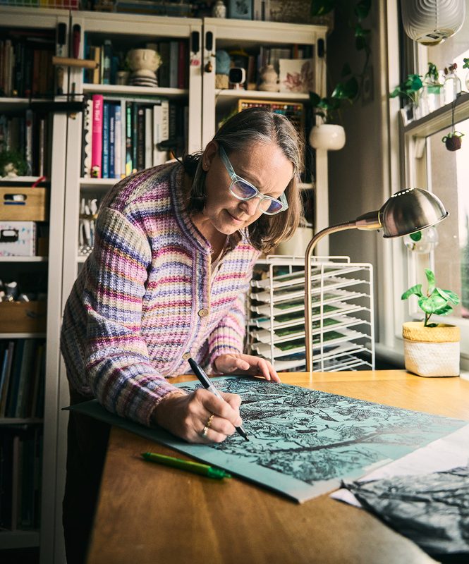 A printmaker in a striped cardigan drawing over a teal print at her desk