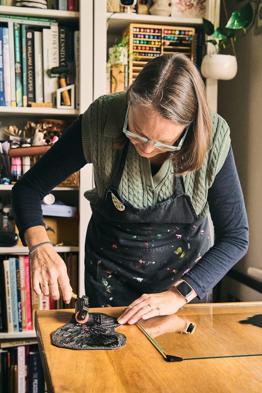 A printmaker in a paint-flecked apron rolling ink across a carved lino block