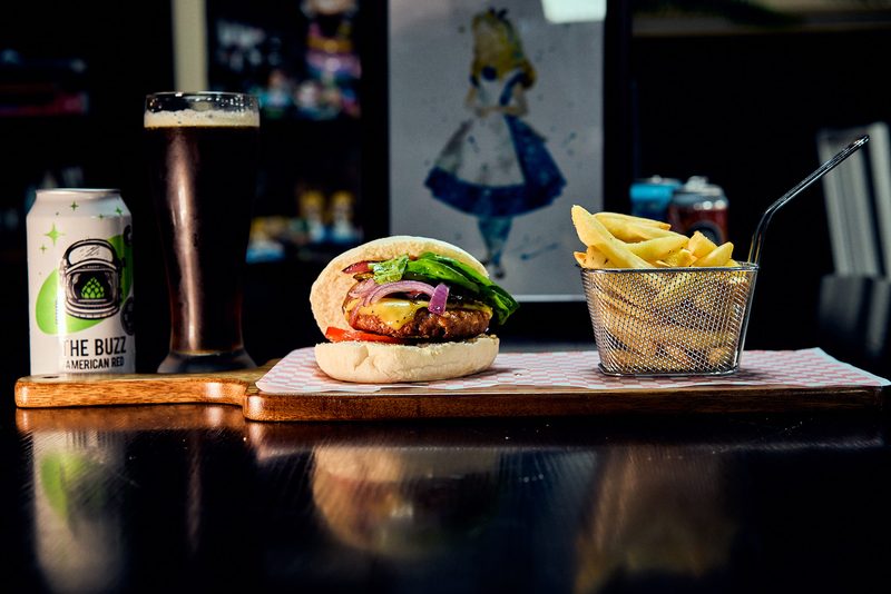 A burger beside fries in a wire basket, with a canned drink and a pint on the bar