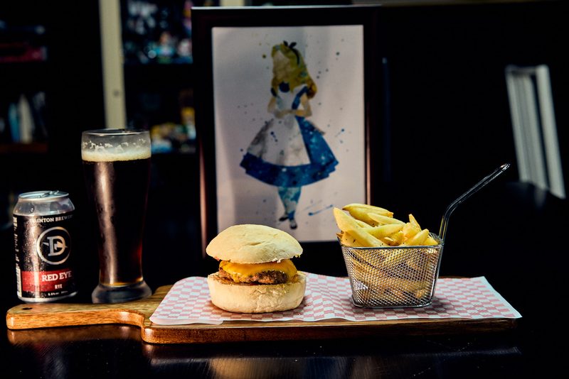 A burger and a basket of fries on a long board, with a can and a pint of dark beer