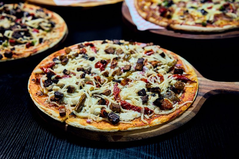 A loaded pizza on a wooden paddle board, more pizzas just out of focus behind it