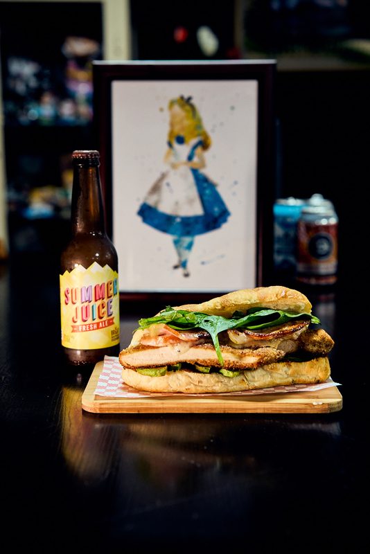A chicken sandwich on a wooden board beside a bottle of juice, an illustrated print in the background