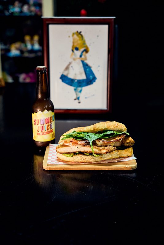 A toasted chicken sandwich on a board with a bottle of juice, under a framed illustration
